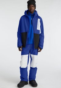 Blue and black snow jacket with white accents, paired with matching blue pants featuring large white patches. Textured fabric and hood.