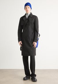 Black double-breasted trench coat, gray zip-up top, black trousers, blue knit beanie, and black chunky shoes, standing against a neutral background.