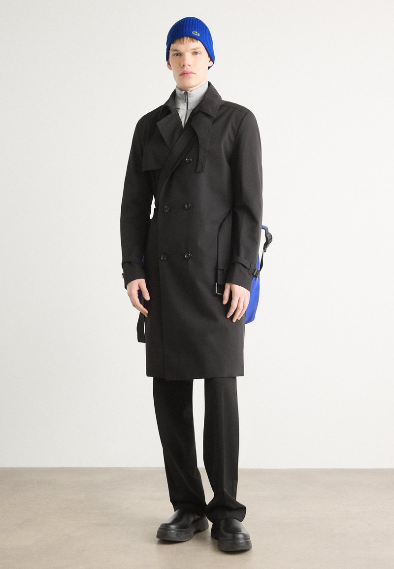 Schwarzer doppelter Trenchcoat, grauer Reißverschluss-Pullover, schwarze Hose, blaue Strickmütze und schwarze klobige Schuhe, die vor einem neutralen Hintergrund stehen.