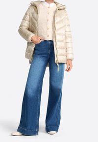 Světle béžová péřová bunda s kapucí, texturovaná tkanina, zapínání na zip; vrstvená přes krémový svetr s kulatými knoflíky; nošena s rozšířenými modrými džíny.