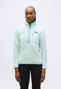 Γυναίκα που φοράει ανοιχτό μπλε fleece pullover της Patagonia με φερμουάρ στην τσέπη στο ύψος του στήθους και μαύρο παντελόνι, στέκεται μπροστά σε απλό λευκό φόντο.