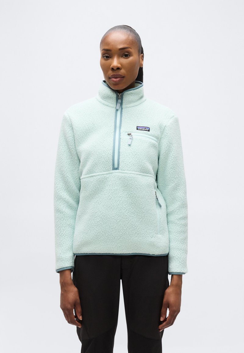 Γυναίκα που φοράει ανοιχτό μπλε fleece pullover της Patagonia με φερμουάρ στην τσέπη στο ύψος του στήθους και μαύρο παντελόνι, στέκεται μπροστά σε απλό λευκό φόντο.