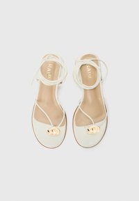 White strappy sandals featuring a smooth leather upper, a gold-tone decorative detail on the toe, and a nude sole with thin laces.