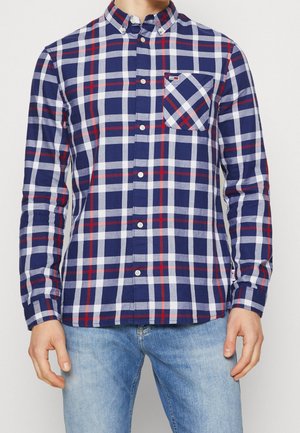 Mann trägt ein blau-weiß-rotes kariertes Hemd mit langen Ärmeln, das zugeknöpft ist und eine Brusttasche hat, kombiniert mit hellblauen Jeans.