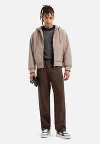 Felpa beige con zip e cordini, indossata sopra un maglione scuro e texturato. Pantaloni marroni e sneakers multicolori completano l'outfit.