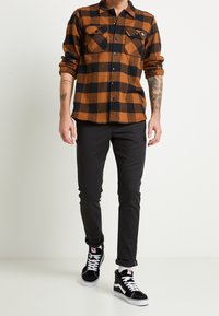 Bruine en zwarte geruite flanellen shirt met twee borstzakken, gecombineerd met zwarte slim-fit broeken en zwarte high-top sneakers met witte accenten.