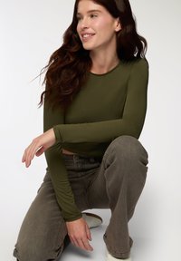 Długi rękaw, oliwkowa bluzka z elastycznego materiału, z okrągłym dekoltem i skróconym końcem, zestawiona z szarymi jeansami z wysokim stanem.