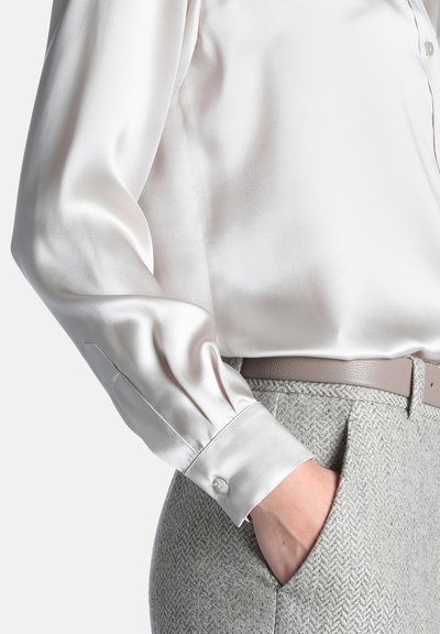 Personne portant un chemisier en satin blanc à manches longues, rentré dans un pantalon en chevrons gris clair avec une ceinture beige, main dans la poche.