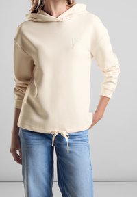 Kremowa bluza z kapturem, wykonana z miękkiego materiału, z logo z przodu, sznurkiem przy hemie oraz luźnym krojem. W zestawie z jasnoniebieskimi dżinsami.