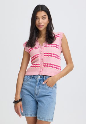 Femme aux longs cheveux foncés portant un haut sans manches en tricot rayé rose et un short en denim bleu clair, posant avec une main dans la poche.