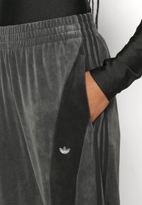 Joggings en velours gris avec accents noirs sur les côtés, taille élastique et poches latérales. Présente un logo argenté en haut à gauche.