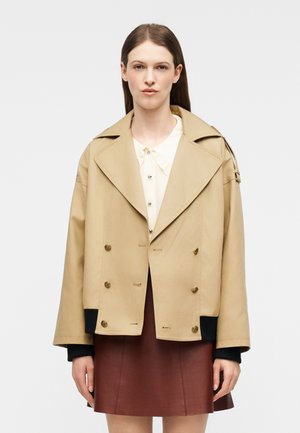 Kobieta ubrana w beżową dwurzędową marynarkę na białej bluzce i brązowej spódnicy, stojąca na jednolitym białym tle.