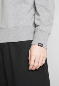 Sudadera gris con puños y dobladillo acanalados. Presenta una pequeña etiqueta negra en el puño. Combinada con pantalones negros. Tejido suave, diseño casual.