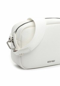 Balta teksturēta plecu soma ar plašu auduma lenti, sudraba rāvējslēdzēju, metāla sprādzi un mazu "SURI FREY" logotipu priekšpusē.