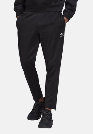 Osoba nosící černé tepláky Adidas s logem na stehně a černé tenisky s vzorovanými tkaničkami, ruce v kapsách.