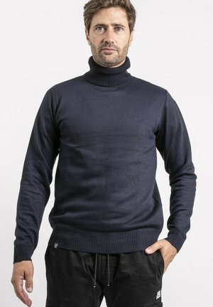 Homme portant un pull à col roulé bleu marine et un pantalon noir, debout contre un fond blanc uni, les mains détendues le long du corps.