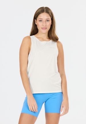 Jeune femme debout portant un haut blanc sans manches et un short bleu vif ajusté devant un fond clair uni.