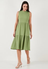 Green midi dress with a round neckline, sleeveless design, textured fabric, and tiered skirt. Worn with nude strappy heels.