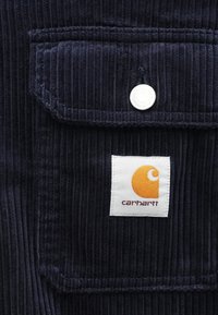 Mörkblå kordflams väv med vertikala ribbor, med en vit etikett med en orange logotyp och en silverfärgad tryckknapp på en bröstficka.
