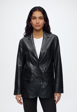 Femme aux longs cheveux foncés portant un blazer en cuir noir par-dessus une chemise blanche et un pantalon noir, debout devant un fond blanc uni.