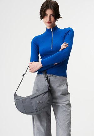 Frau trägt ein blaues Oberteil mit Reißverschluss und graue Hose, hält eine große graue Handtasche mit verstellbarem Schultergurt.