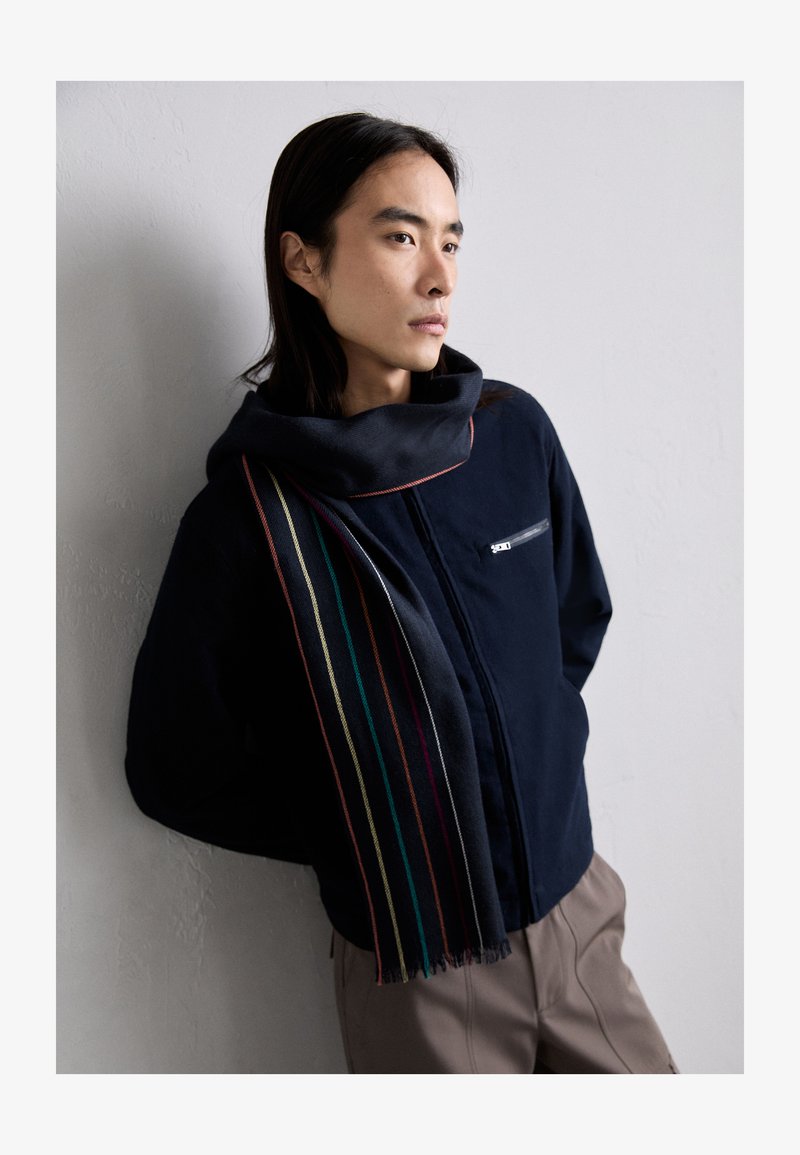 Jūras spēku zips-jaka ar augstu apkakli un krūškurvja kabatu, apvienota ar tumšu šalli, kurai ir vertikāli daudzkrāsaini svītri.