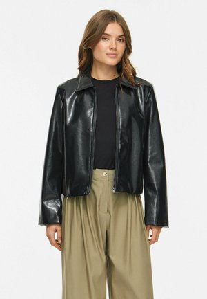 Femme portant une veste noire brillante zippée sur un haut noir et un pantalon beige taille haute à jambes larges, debout devant un fond blanc uni.