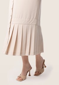 Jupe plissée beige avec un devant à boutons et une taille ajustée, associée à des mules à talons ouvertes couleur nude avec un design à bout carré.