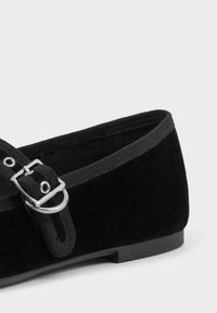Scarpa piatta in suede nera con punta arrotondata, accentuata da una fibbia decorativa e una finitura liscia e opaca. Tacco basso e rettangolare.