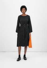 Svart kjole med blomstermønster, belted midje og lange ermer. Modellen holder en oransje veske og har på seg svarte sokker og loafers.