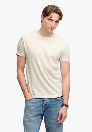 Joven con cabello ondulado que lleva una camiseta de manga corta color beige claro, vaqueros azules y un reloj de pulsera oscuro sobre fondo blanco.