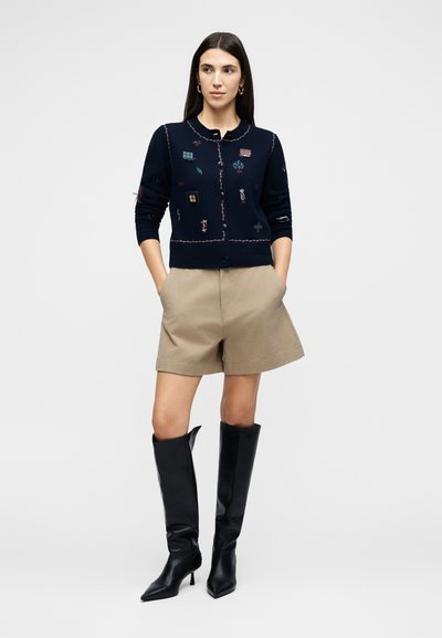 Γυναίκα που φοράει μπλε cardigan με πολύχρωμα μπαλώματα, μπεζ σορτς και μαύρα μπότες μέχρι το γόνατο με τακούνια, στέκεται με τα χέρια στις τσέπες.