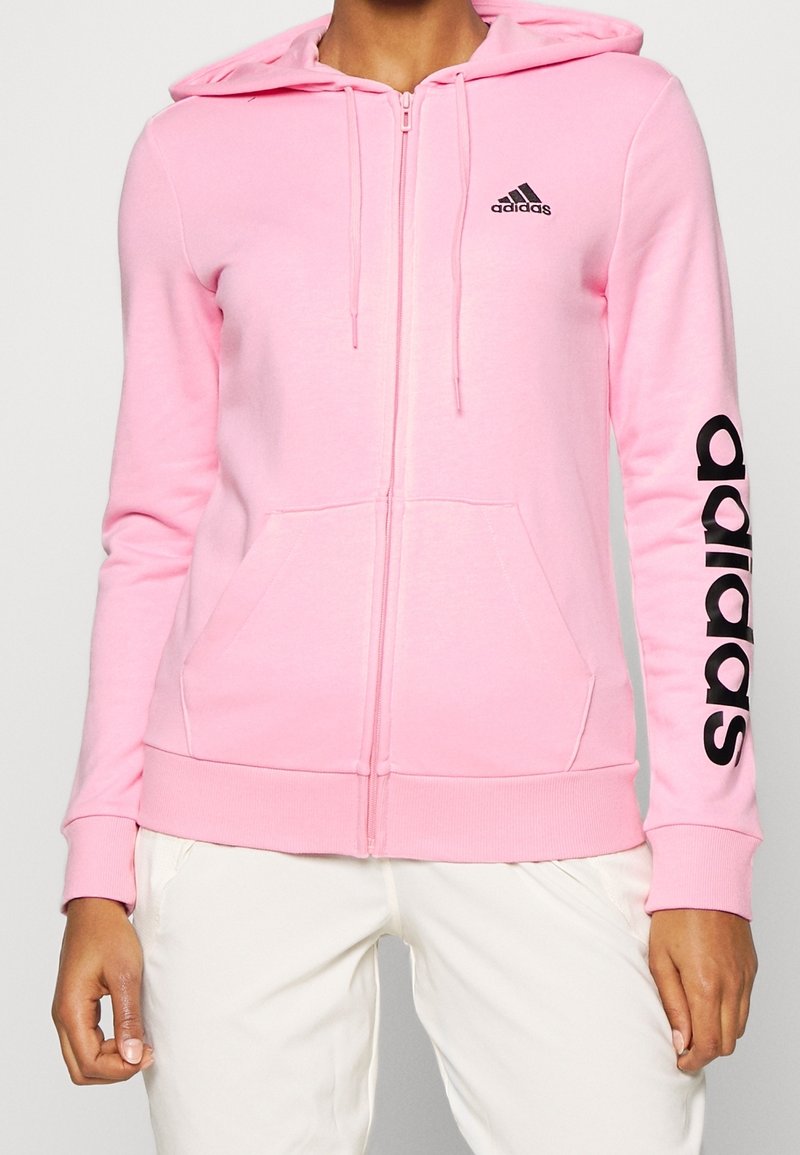 Sudadera rosa con cremallera, fabricada en un tejido suave. Cuenta con una capucha, bolsillos frontales y el logotipo negro de "adidas" en el lado izquierdo del pecho y a lo largo de la manga.