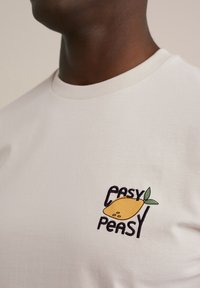 Valge puuvillane t-särk, millel on tikandiga muster, kus on sõnad "EASY PEASY" ja kollane sidrun, millele on lisatud rohelised lehed.