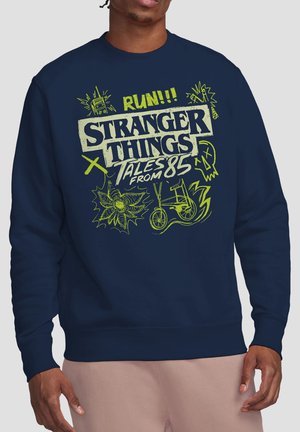 Navy sweatshirt with neon green Stranger Things text, illustrations of a radio, bike, skull, and spider-like creature, worn by a man.