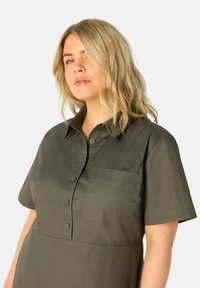 Camicia a maniche corte con bottoni in tessuto verde oliva scuro, dotata di taschino sul petto e texture liscia con collare strutturato.