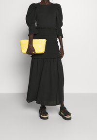 Top nero con maniche a palloncino e gonna maxi a più strati con dettagli smock, abbinata a una clutch gialla intrecciata e sandali neri con zeppa e frange.