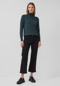 Femme portant un pull à col roulé vert foncé, un jean noir court avec un ourlet effiloché et des mocassins noirs épais sur un fond uni.