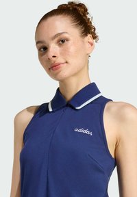 Mladá žena s červenými kučeravými vlasmi v drdole, oblečená v bezrukávovom námořníckom modrom polo tričku Adidas s bielym límcom.