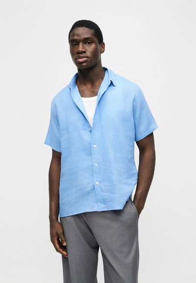 Άνδρας με σκουρόχρωμο δέρμα που φοράει ανοιχτό μπλε κοντομάνικο πουκάμισο πάνω από λευκό μπλουζάκι και γκρι παντελόνι, στέκεται μπροστά σε απλό φόντο.