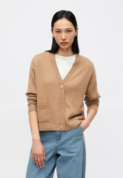 Cardigan en laine beige avec un col en V côtelé, boutonné sur le devant, et deux poches avant. Associé à un jean en denim bleu clair. Texture lisse.
