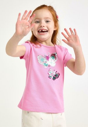 Ung rød-håret pige, der smiler og vinker med hænderne. Hun har en lyserød t-shirt på med to paillet-dekorerede sommerfugle og lyse bukser.