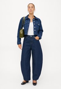 Donkerblauwe spijkerjas met knoopsluiting en borstzakken, gecombineerd met wijdvallende donkerblauwe jeans. Model houdt een groene tas vast.
