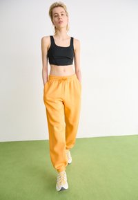 Top corto negro combinado con pantalones deportivos naranjas, que cuentan con una cinturilla elástica y bolsillos laterales. Los zapatos son de color gris claro con acentos amarillos.