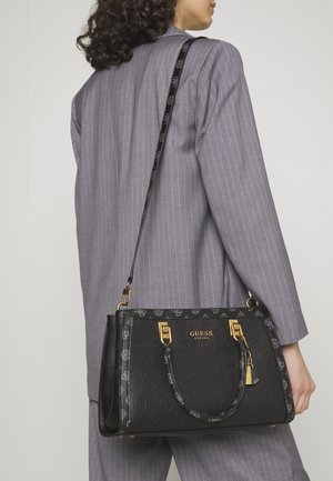 Person in grauem Nadelstreifenanzug trägt eine schwarze GUESS-Handtasche mit goldener Hardware und Logo auf der Schulter.