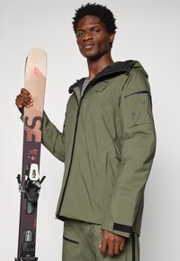 Zöld vízálló dzseki kapucnival, cipzárakkal és texturált anyaggal; hódeszkát tart, fekete kötésekkel és bézs, piros díszítéssel a tetején.