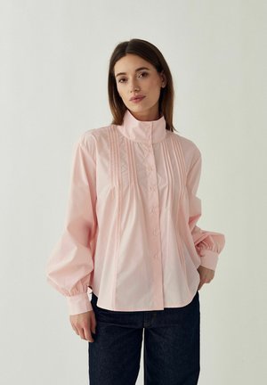 Kvinde iført en lyserød bluse med knapper, vertikale plisseringer og puffede lange ærmer, kombineret med mørkeblå jeans mod en ensfarvet baggrund.