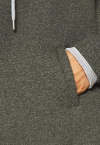 Dunkelgrüne Kapuzenjacke aus einem strukturierten Stoff, mit einer Fronttasche und grauen Rippstrickbündchen. Mit einem Kordelzug am Kragen.