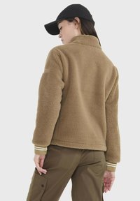Braune Fleecejacke mit hohem Kragen, cremefarbigen und olivgrünen gestreiften Bündchen sowie einer lockeren Passform. Über olivgrünen Hosen und einer schwarzen Mütze getragen.