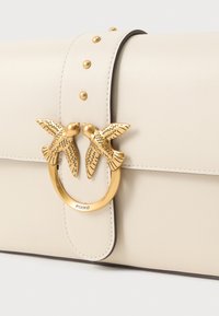 Beige bőr kézitáska arany madár alakú záródással, kerek díszítéssel és szegecses részletekkel a fedélen. Sima felület, kontrasztos varrással.